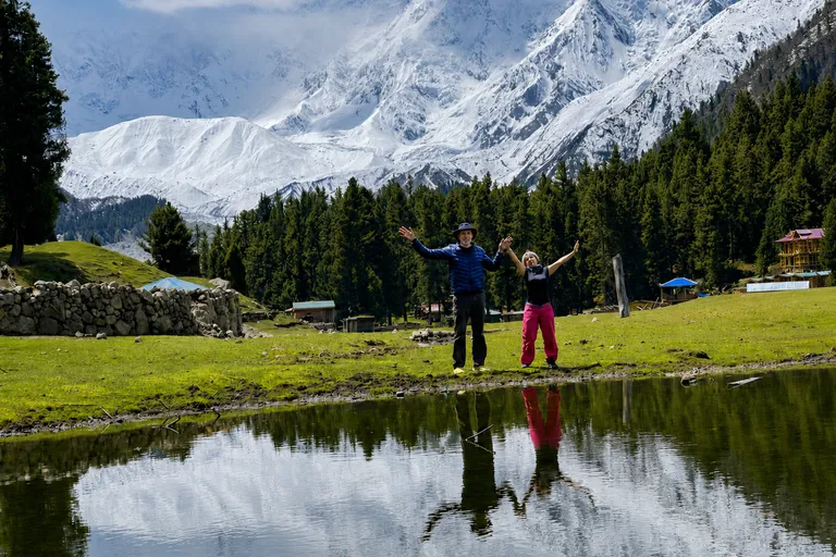 Two travellers with arms raised on the green Fairy Meadows alpine meadow, with the snow face of Nanga Parbat behind and a pond reflection in front