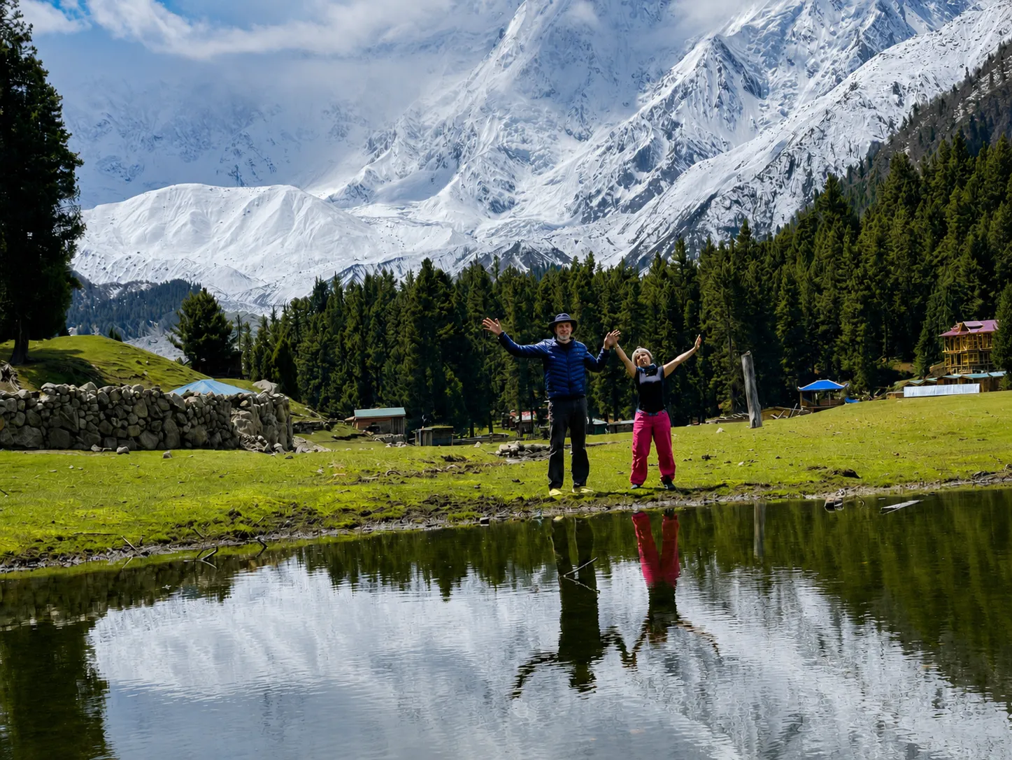 Two travellers with arms raised on the green Fairy Meadows alpine meadow, with the snow face of Nanga Parbat behind and a pond reflection in front