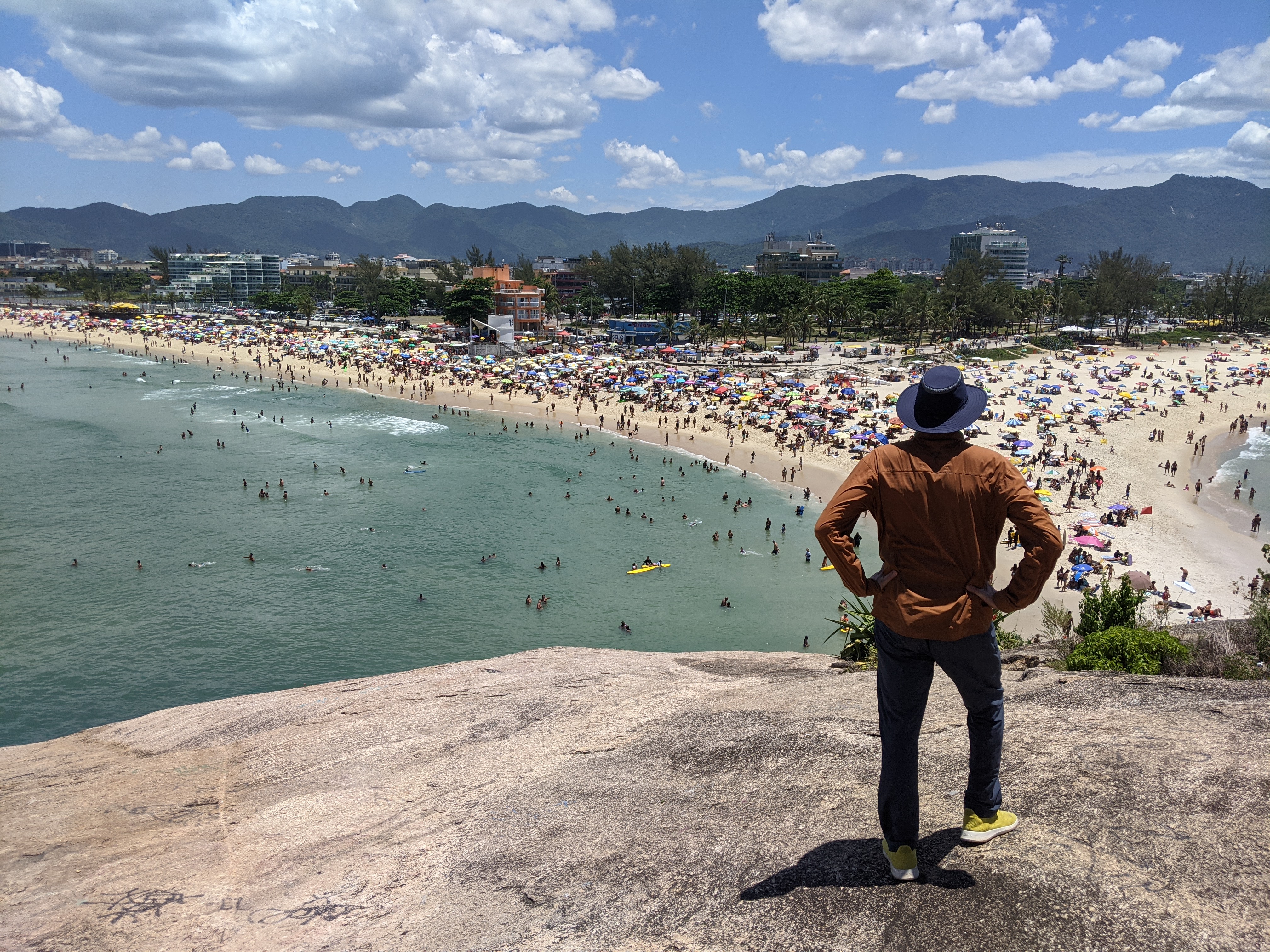 Traveller standing on a rock above a busy beach with green water and mountains near Rio de Janeiro