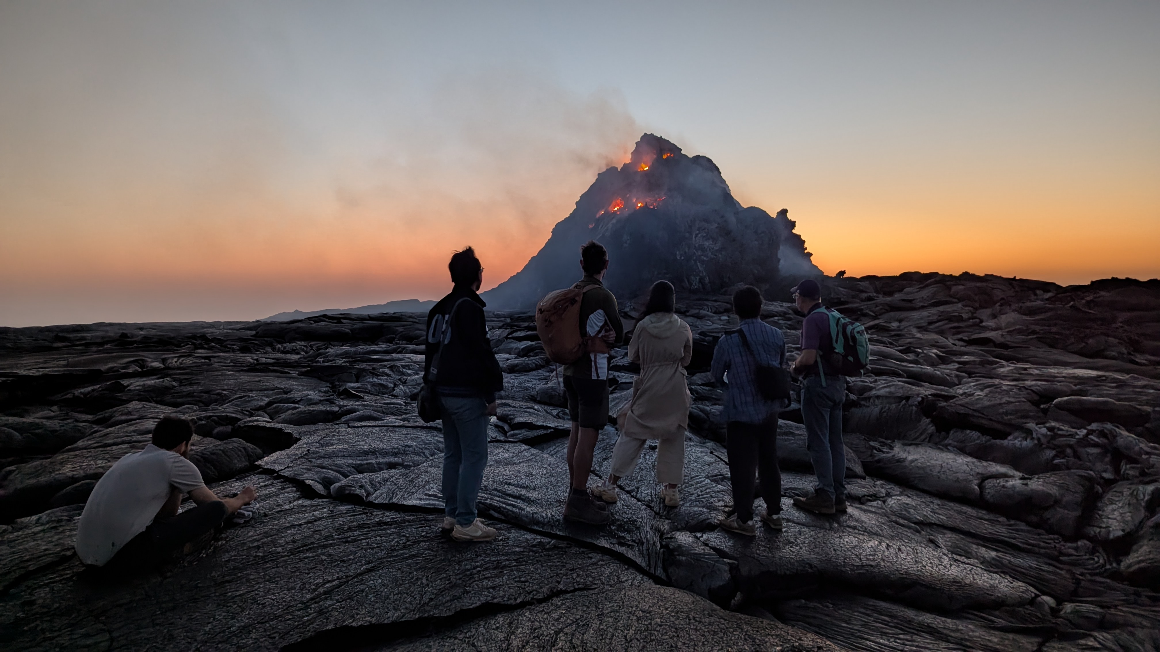 Group standing on cooled black lava watching the glowing Erta Ale lava cone at dusk