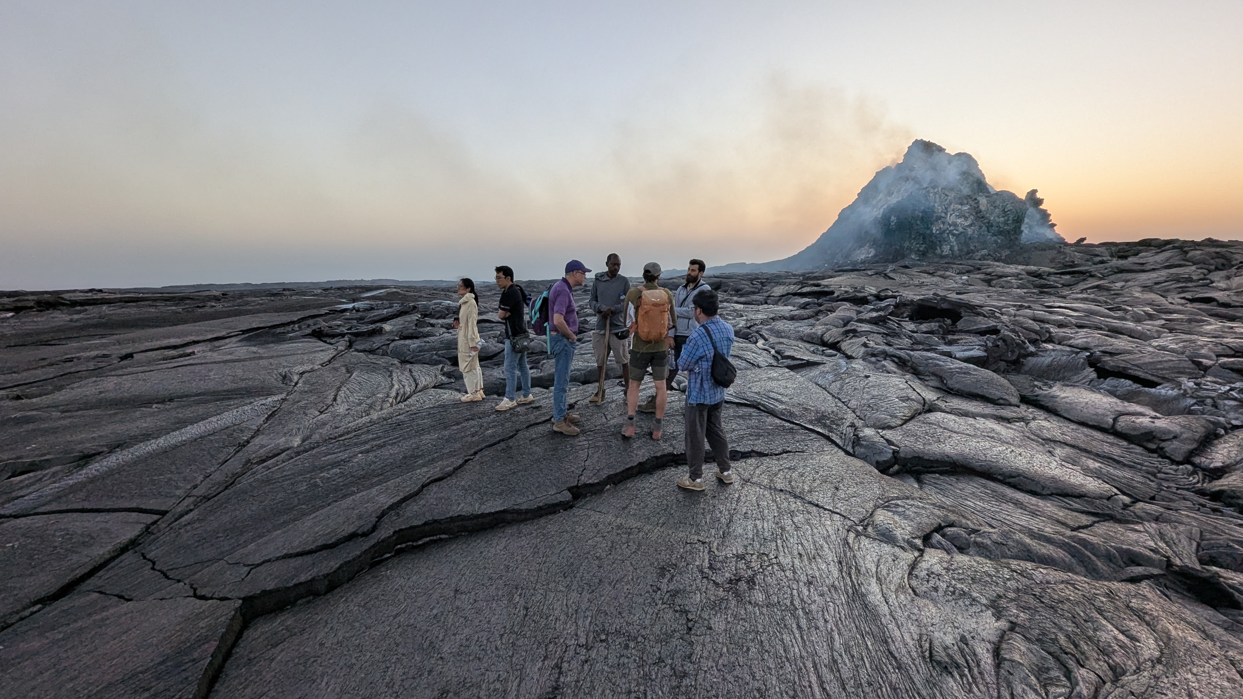 Group on a cracked black lava field with the smoking Erta Ale cone behind at dawn