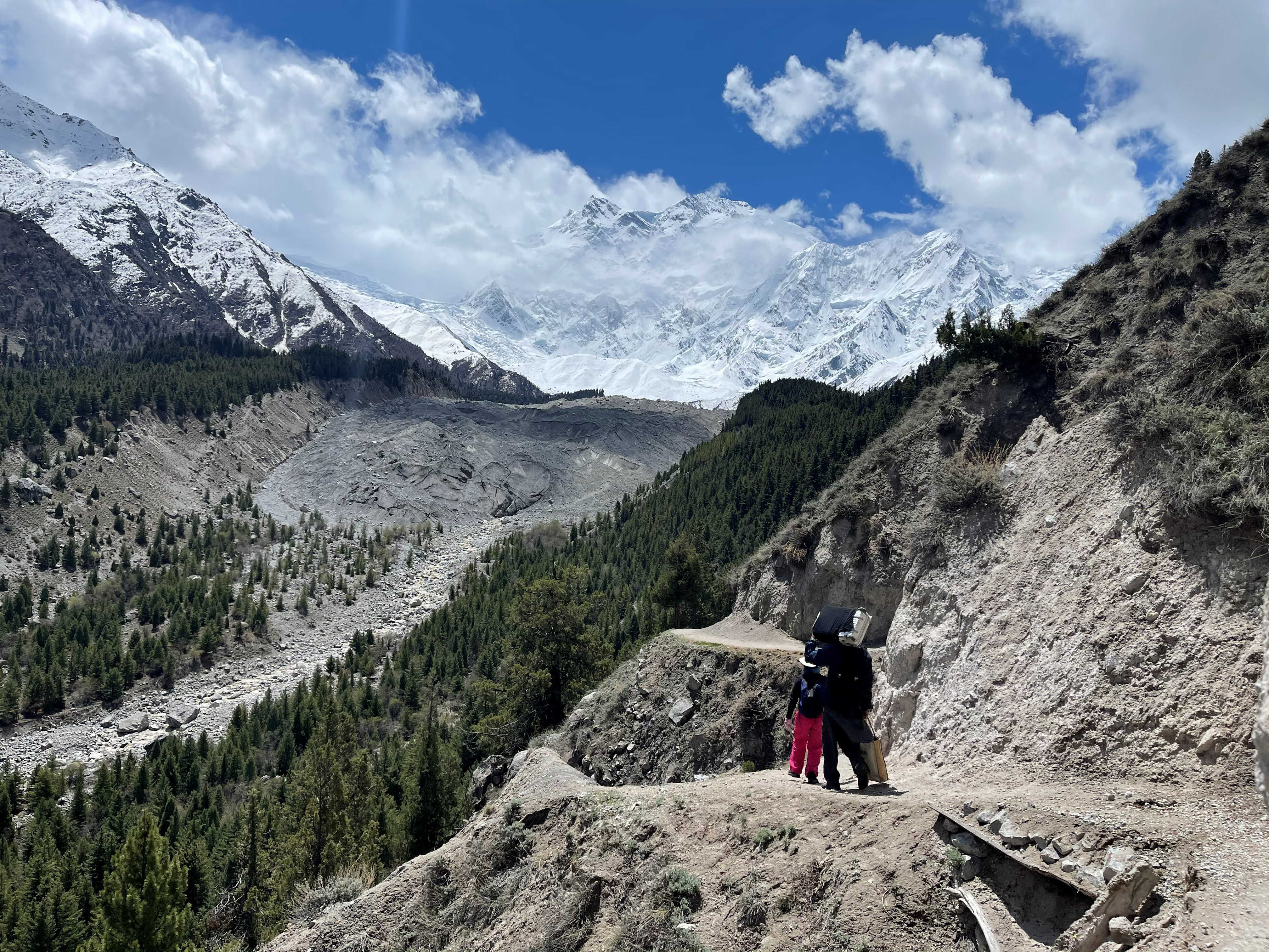 Travellers on a dusty mountain trail looking toward the snow-covered face of Nanga Parbat across a glacial valley