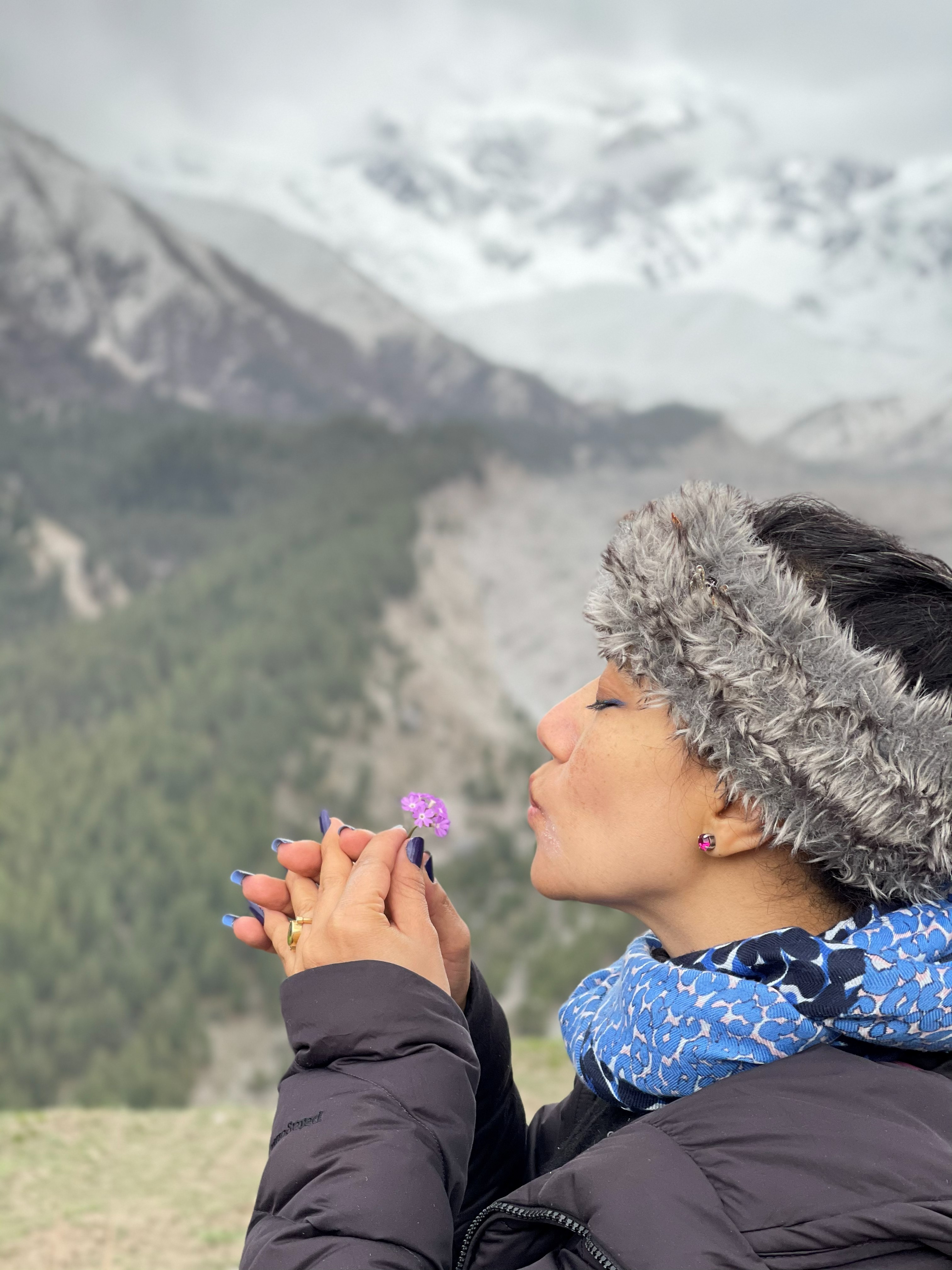 Traveller in a fur hat and down jacket holding a small purple wildflower with misty mountains behind