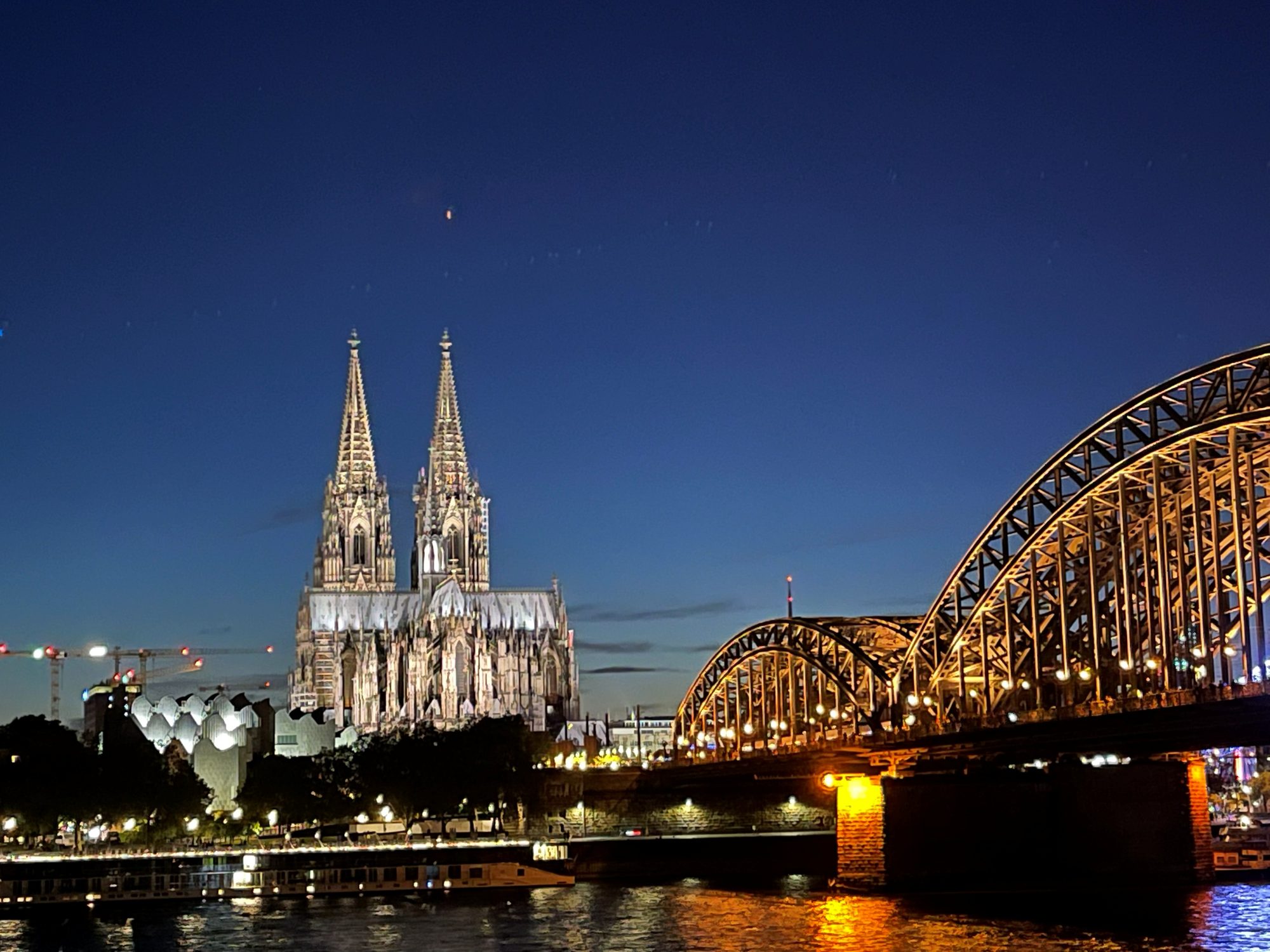 Cologne Cathedral floodlit at night beside the illuminated Hohenzollern Bridge, reflected in the Rhine