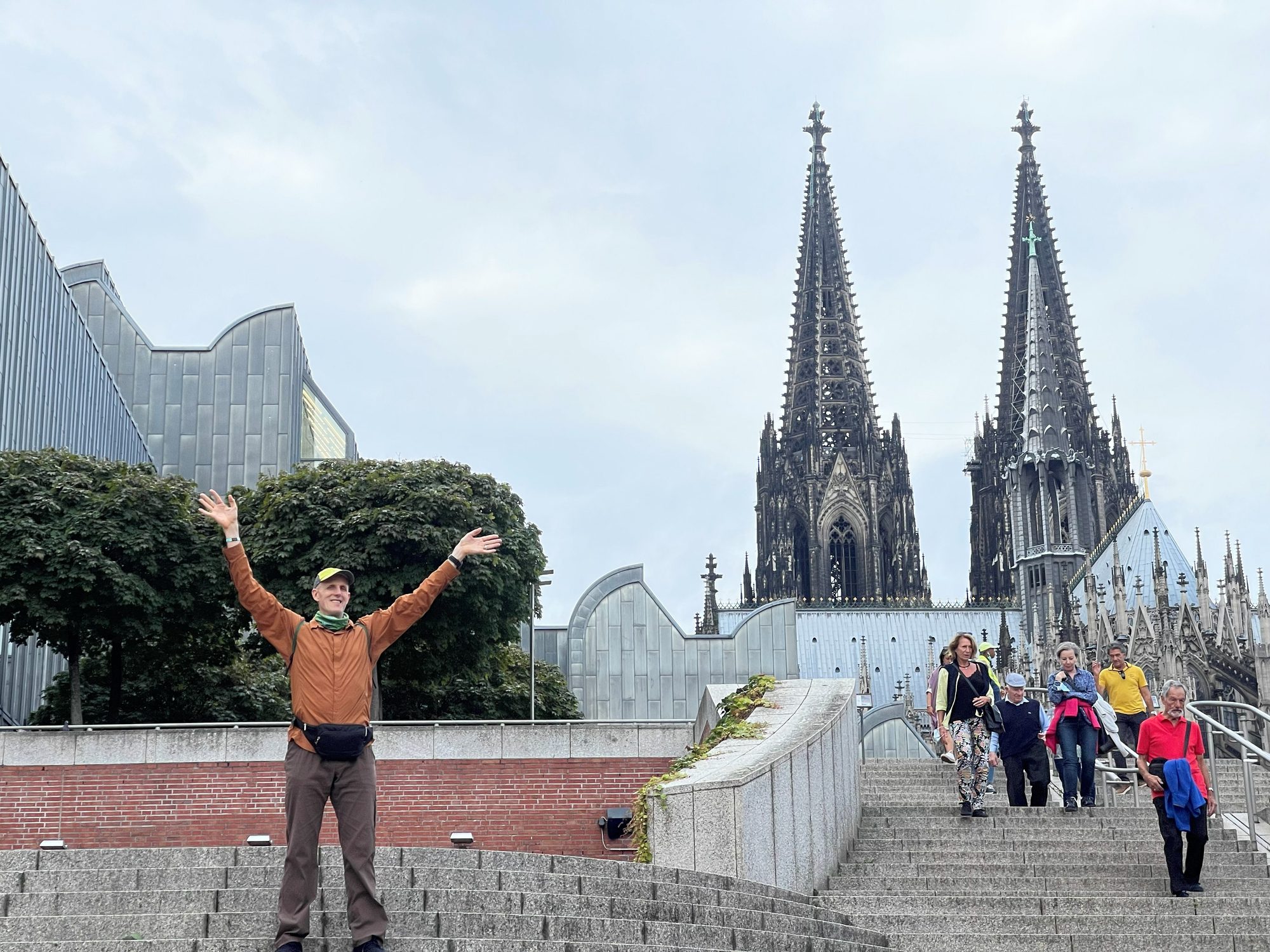 Traveller with arms raised on steps below Cologne Cathedral's twin spires, with the Museum Ludwig alongside
