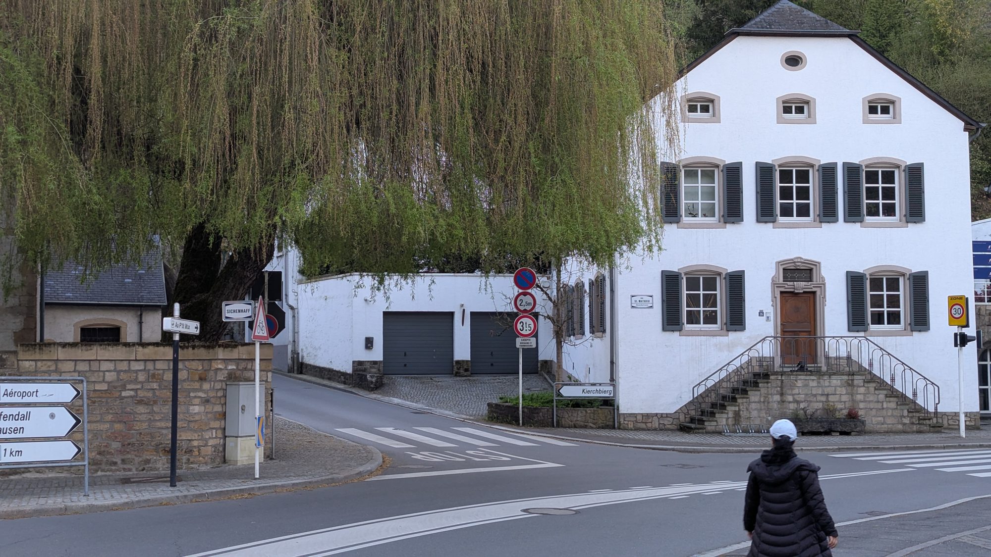 Whitewashed Luxembourgish house with green shutters beside a large weeping willow at a village road junction