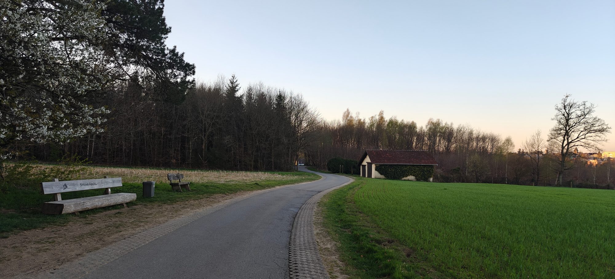 Quiet country road at dusk with a bench, a small stone hut, fields and forest in rural Luxembourg