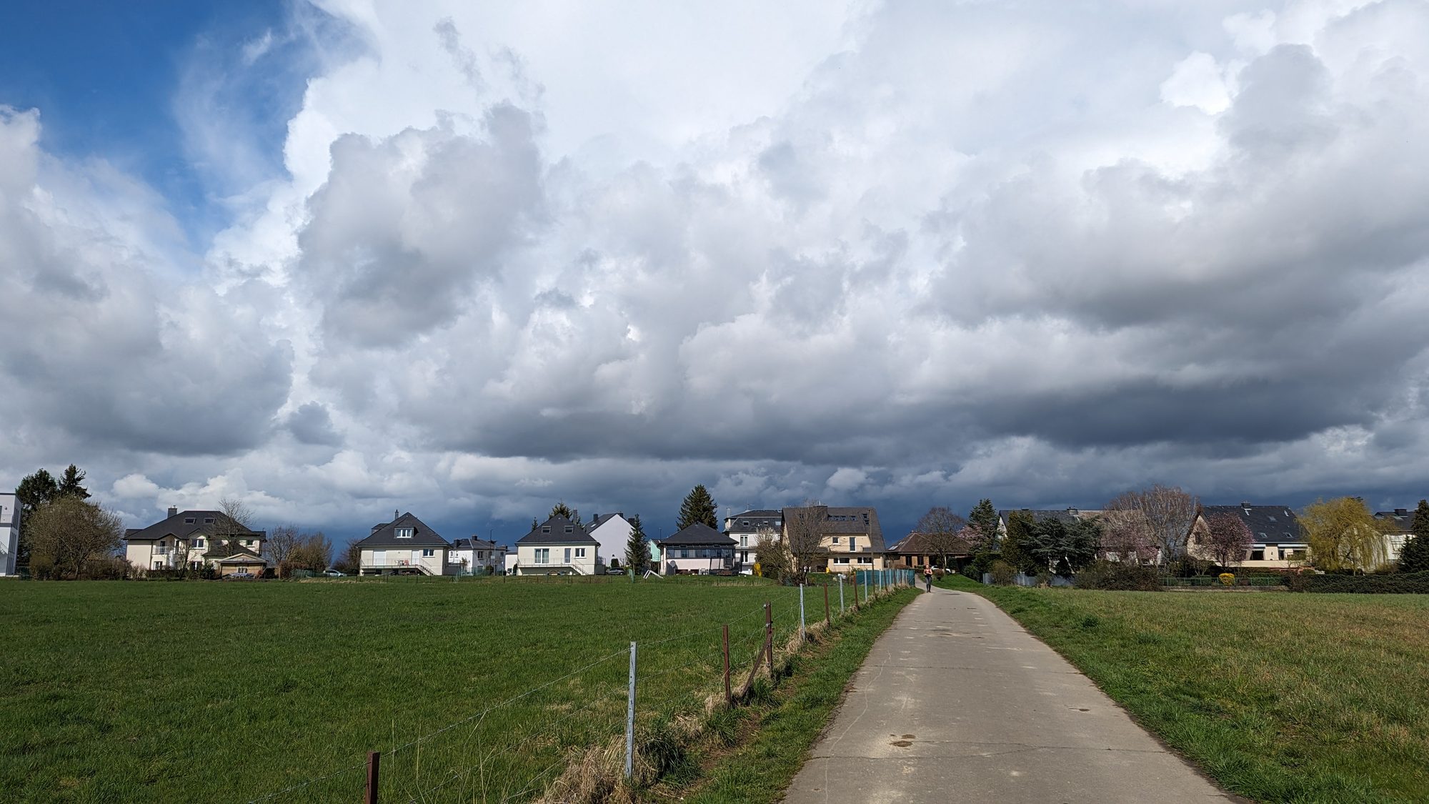 Dramatic cloudy sky over a path between green fields with a row of suburban houses in Luxembourg
