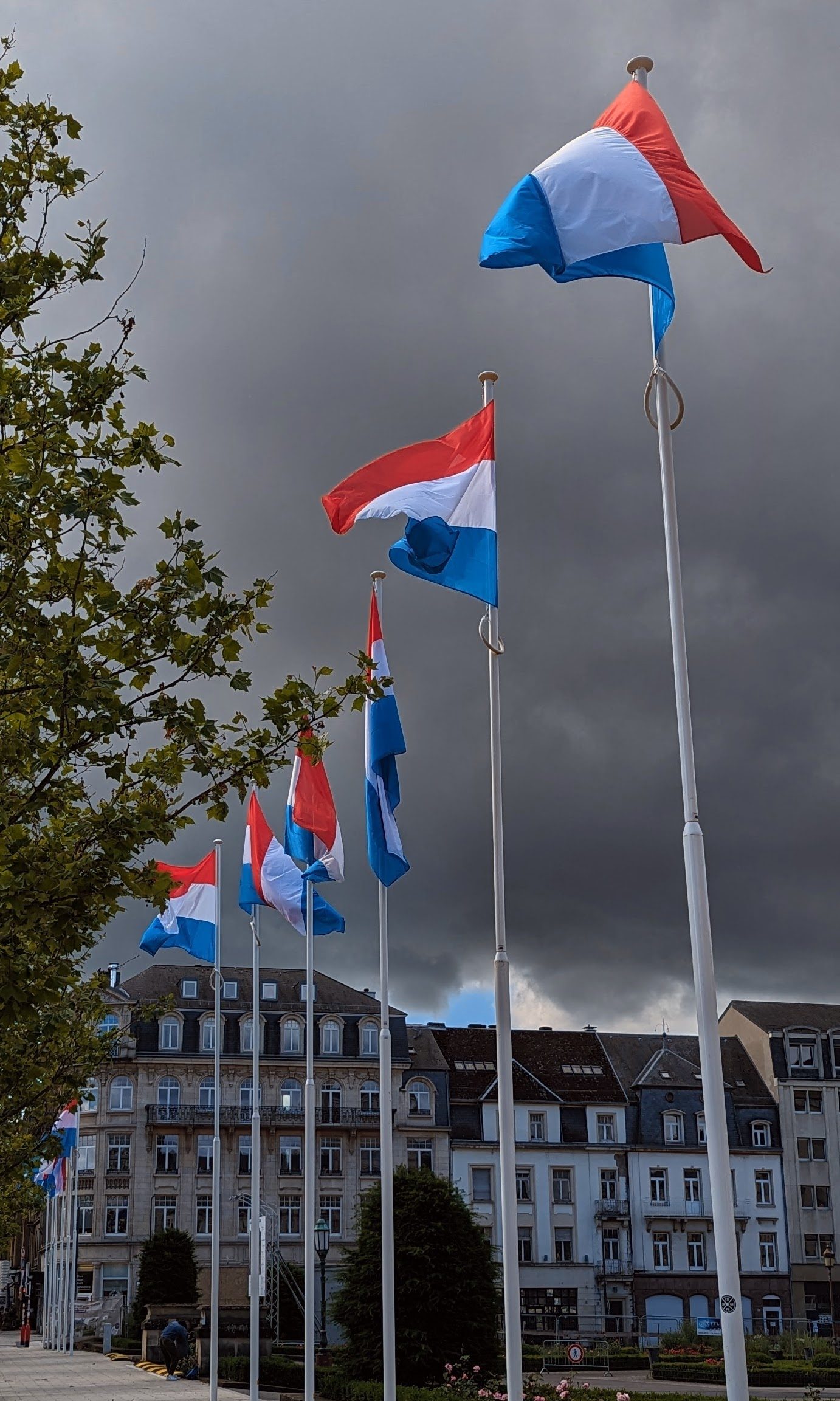Red, white and blue Luxembourg national flags flying on poles in a city square under a grey sky